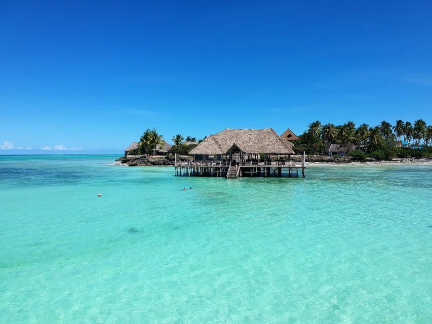 Una capanna di legno con tetto di paglia su palafitte si erge su acque limpide e turchesi, con una costa tropicale e fiancheggiata da palme sullo sfondo.