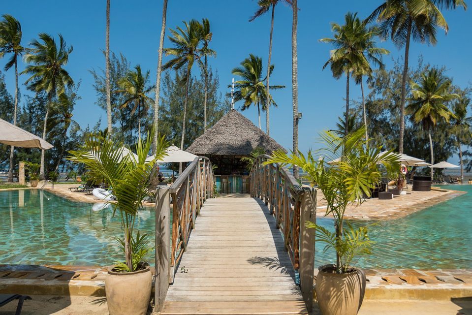 Un ponte di legno attraversa la piscina di un resort fino a un bar con tetto di paglia, circondato da alte palme.