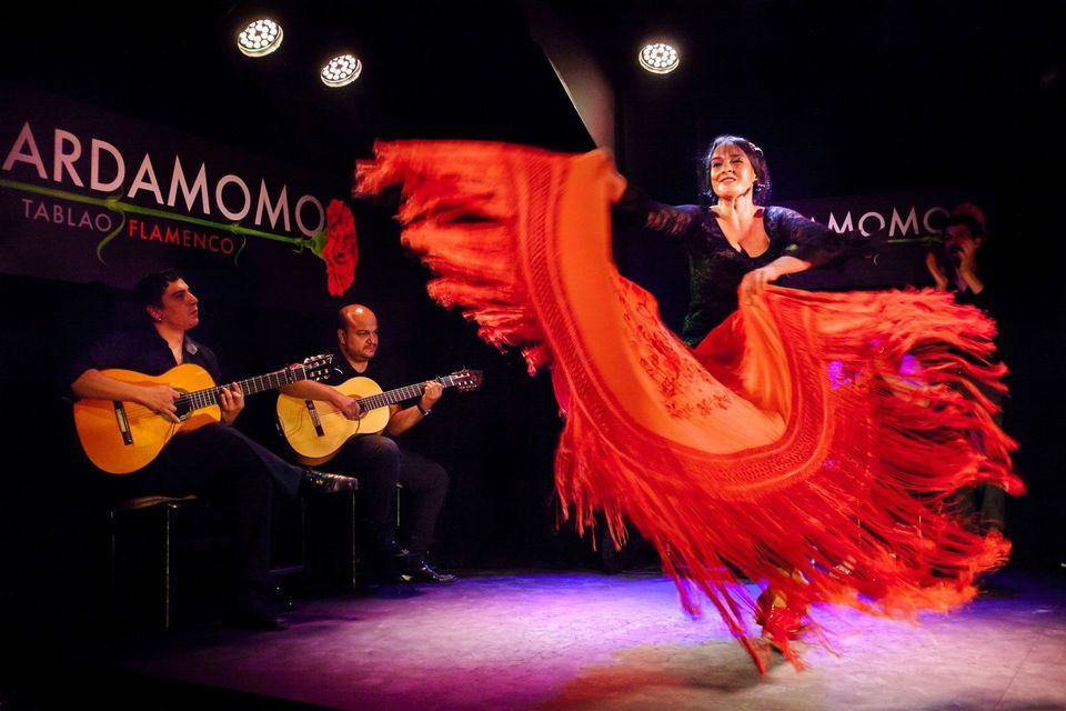 Una ballerina di flamenco fa roteare un grande scialle rosso frangiato su un palcoscenico, accompagnata da due chitarristi che suonano sotto i riflettori.