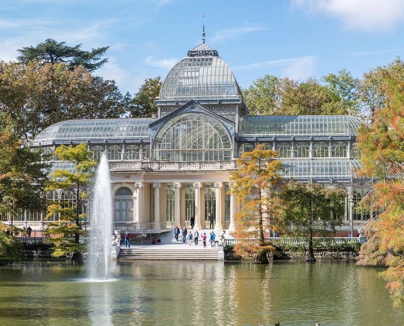 Un sontuoso palazzo di vetro con una cupola si erge dietro un laghetto con una grande fontana, circondato da alberi in un parco.