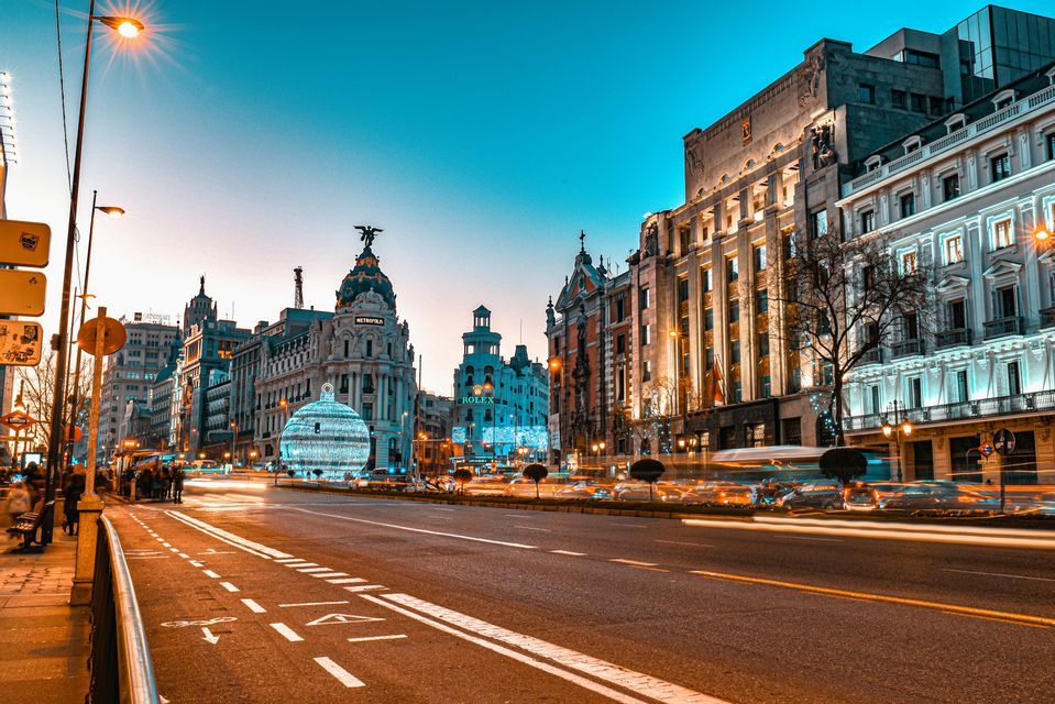 Un'ampia strada cittadina al crepuscolo, con palazzi ornati e illuminati e scie luminose del traffico in lunga esposizione.