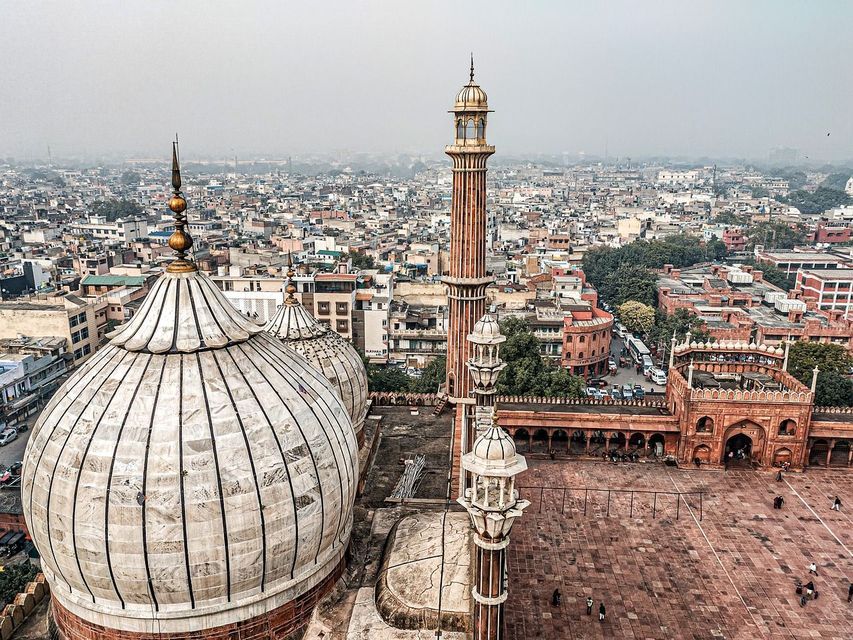 Vista aérea de las cúpulas blancas y el alto minarete de una mezquita sobre un extenso y brumoso paisaje urbano.