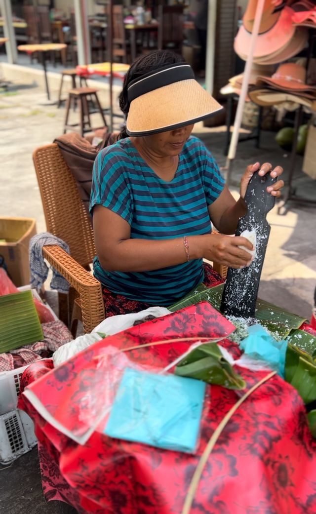Una donna con una camicia a righe e una visiera di paglia grattugia il cocco su una foglia di banana mentre è seduta a una bancarella del mercato all'aperto.