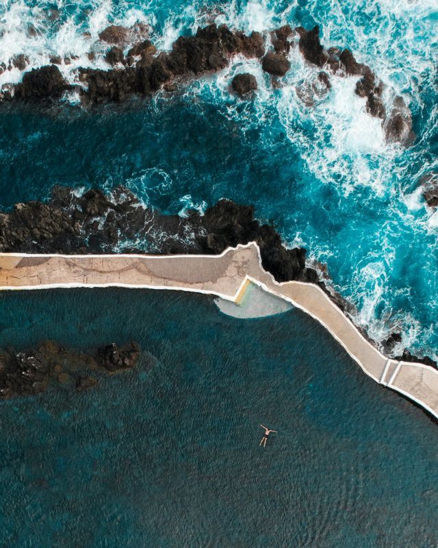 Una vista aerea di una persona che galleggia in una calma piscina naturale accanto all'oceano agitato e rocce scure.