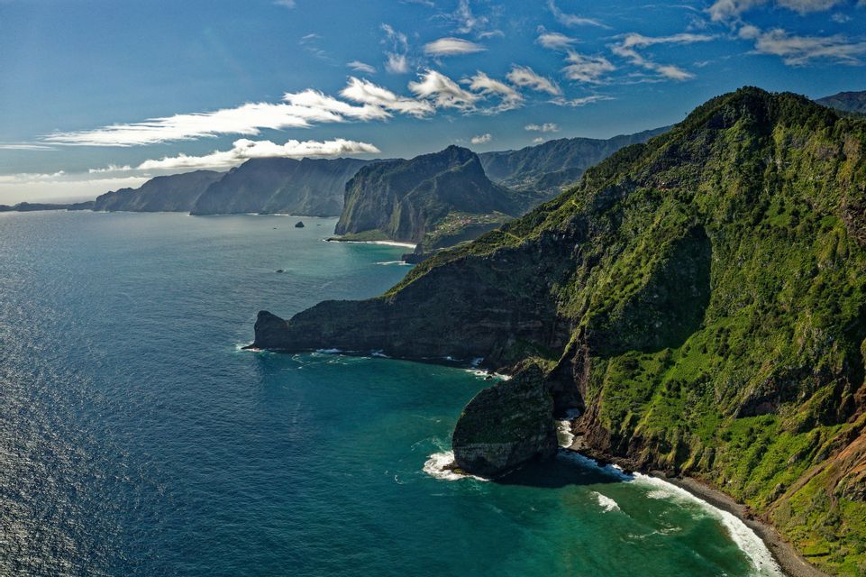 Ripide scogliere verdi si tuffano nell'oceano blu lungo una costa frastagliata sotto un cielo soleggiato con nuvole bianche.