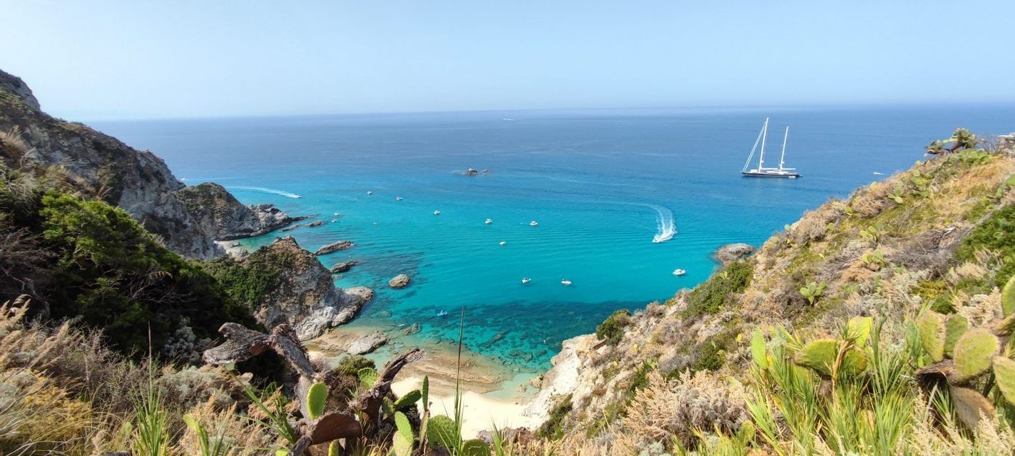 Vista dall'alto di una costa rocciosa con una spiaggia appartata e acqua turchese, dove diverse barche e una grande barca a vela fluttuano sotto un cielo limpido.
