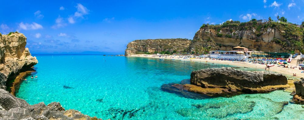 Una vista panoramica di una cala sabbiosa con acqua turchese, incorniciata da grandi scogliere sotto un cielo azzurro chiaro.