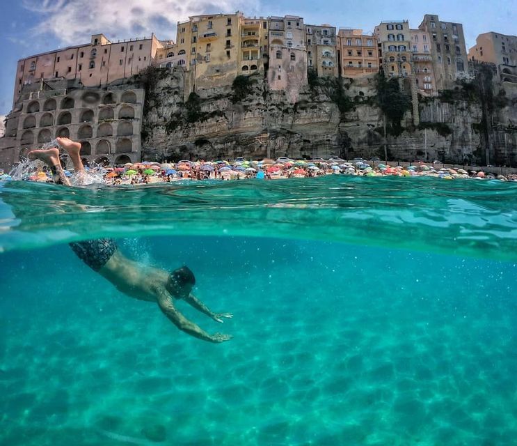 Una foto split-shot di una persona che nuota sott'acqua nel mare turchese, con una spiaggia affollata e una città sulla scogliera sopra.