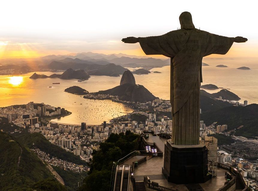 Blick von hinter einer großen Statue mit ausgebreiteten Armen, die eine weitläufige Stadt, Bucht und Berge bei Sonnenuntergang überblickt.