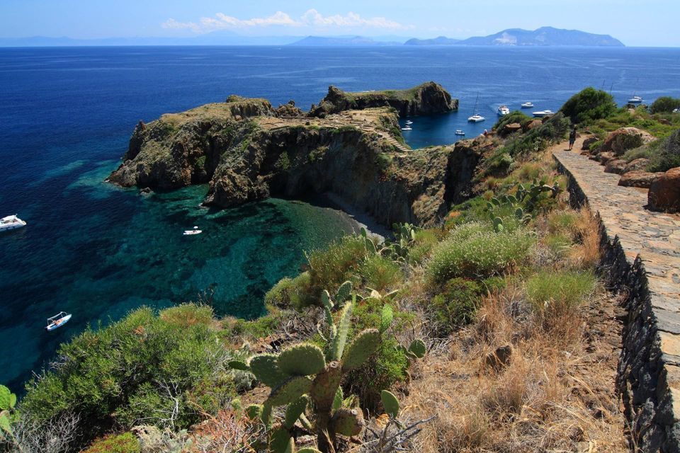 Una vista costiera soleggiata da un sentiero in pietra, che si affaccia su una caletta rocciosa con acqua turchese e diverse barche ancorate.