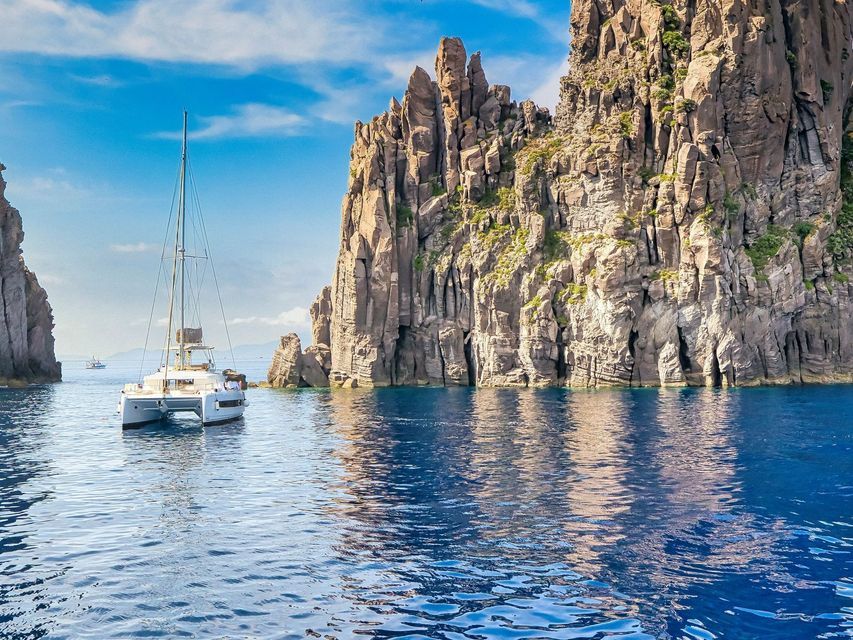 Un catamarano bianco galleggia su acque calme e blu, accanto a una scogliera rocciosa a picco, sotto un cielo azzurro.