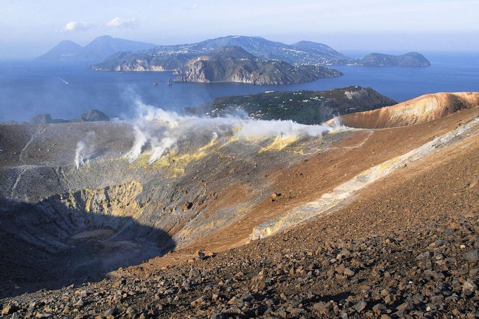 Fumo si alza da un cratere vulcanico con pendii rocciosi, offrendo una vista di un'isola lontana attraverso il mare blu.
