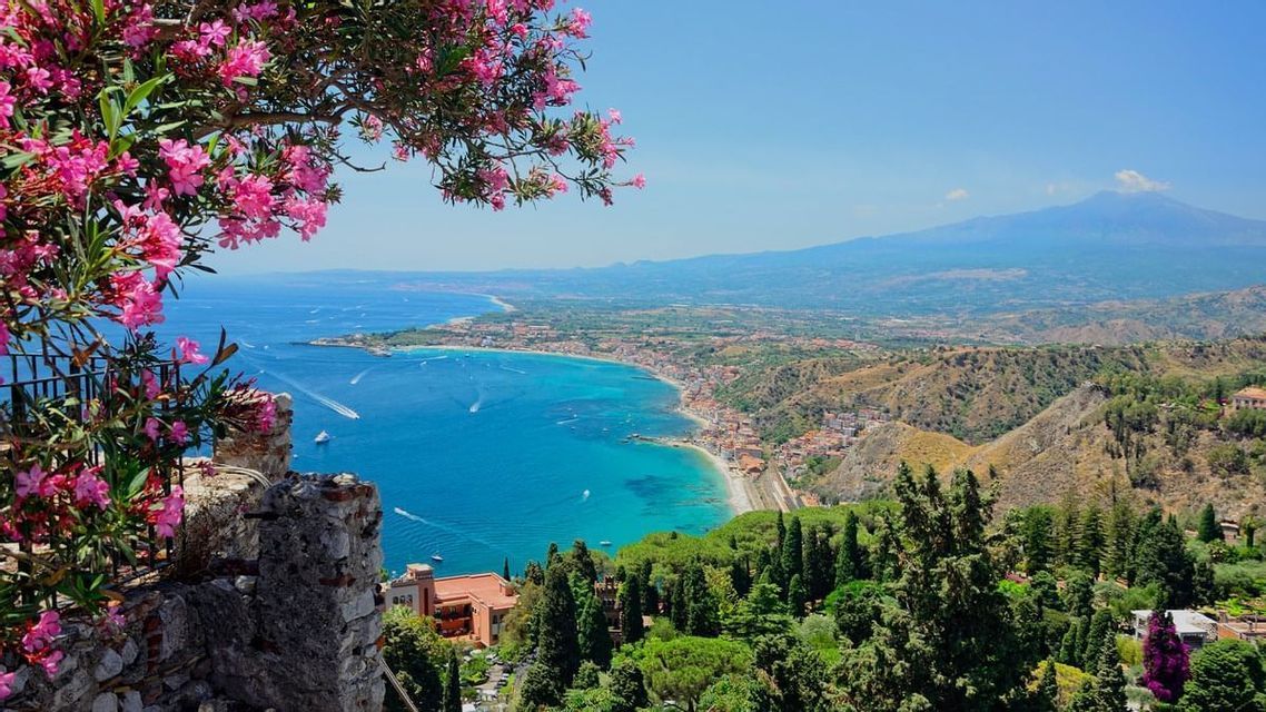 Flores rosas en un árbol enmarcan una vista elevada de un pueblo costero, una bahía turquesa y una montaña lejana bajo un cielo azul claro.