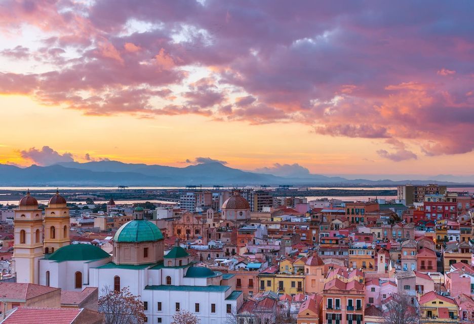 Un paesaggio urbano di edifici colorati e cupole storiche visto al tramonto, con montagne e uno specchio d'acqua sullo sfondo.
