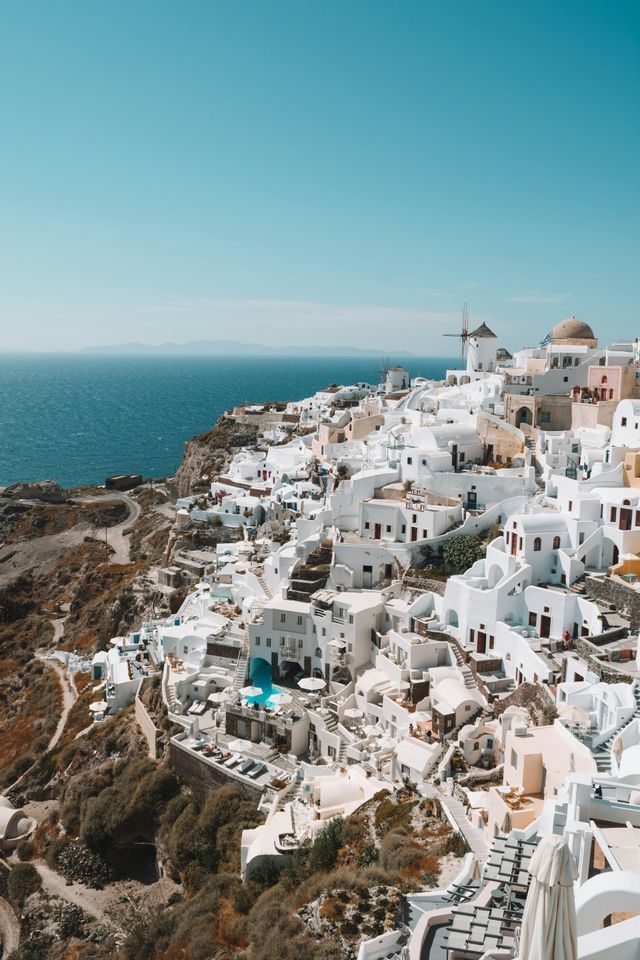 Un villaggio di edifici imbiancati a calce e mulini a vento arroccato su una scogliera che domina un mare blu calmo sotto un cielo sereno.