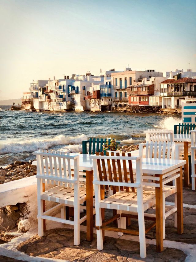 Tavoli e sedie bianchi su un patio in pietra si affacciano sulle onde che si infrangono sulla costa di una città costiera.