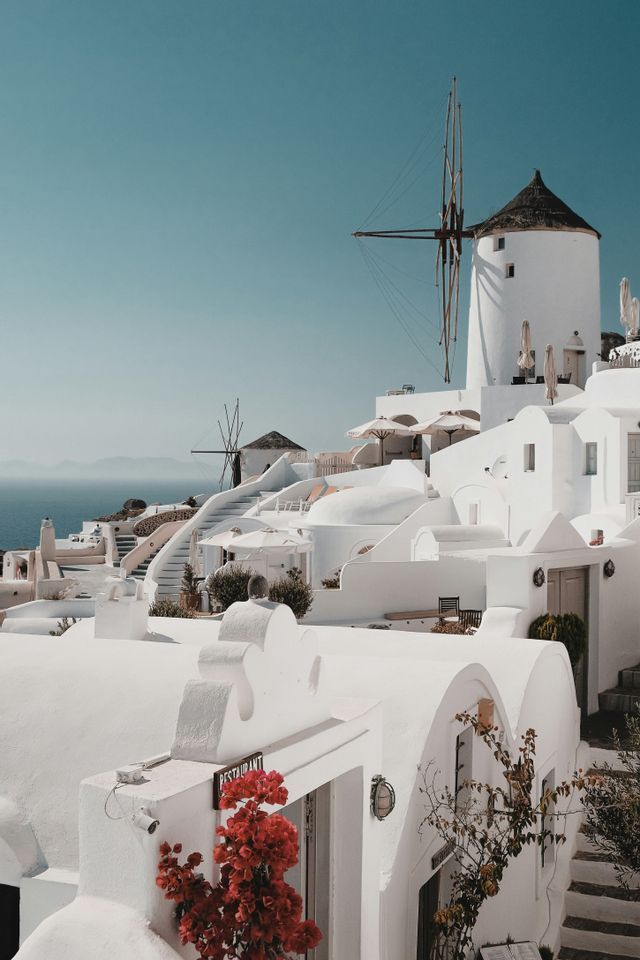 Edifici imbiancati a calce e mulini a vento tradizionali su una collina che domina il mare sotto un cielo sereno.