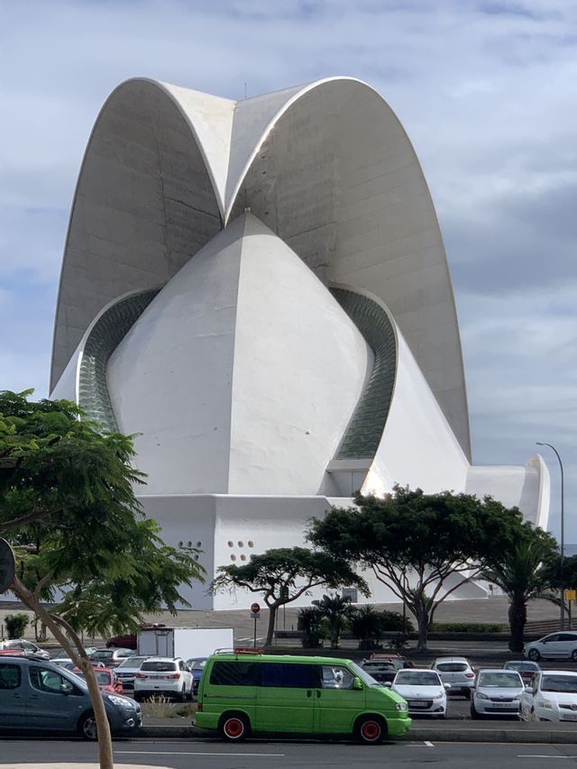 Un gran edificio blanco, con un techo ancho y curvo, se encuentra detrás de una calle bordeada de autos estacionados, incluida una camioneta verde brillante.