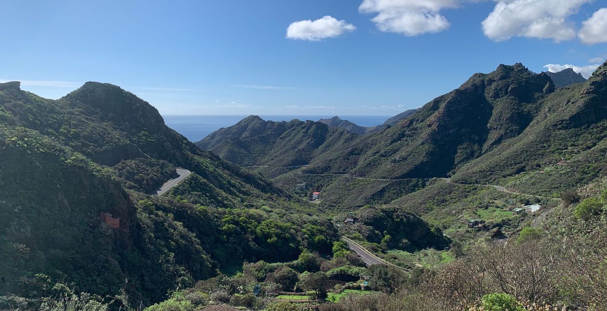 Un camino sinuoso serpentea a través de un exuberante valle verde de montaña con el océano visible en la distancia bajo un cielo azul con nubes blancas.