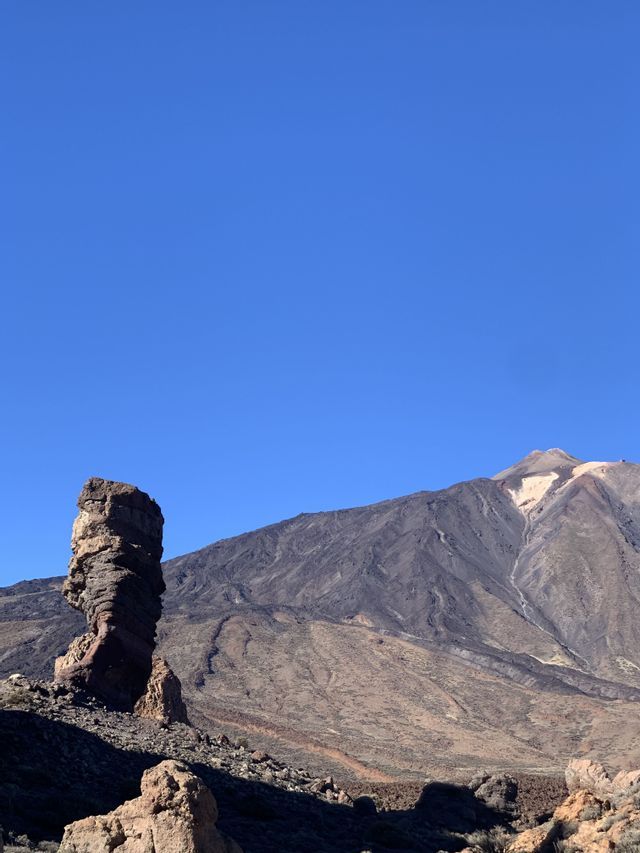Una gran formación rocosa estratificada se eleva en un paisaje volcánico con una imponente montaña detrás bajo un cielo azul claro.