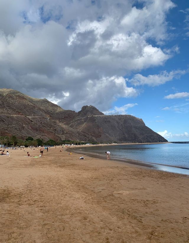 Gente disfrutando de un día en una amplia playa de arena al pie de una gran montaña rocosa bajo un cielo nublado.