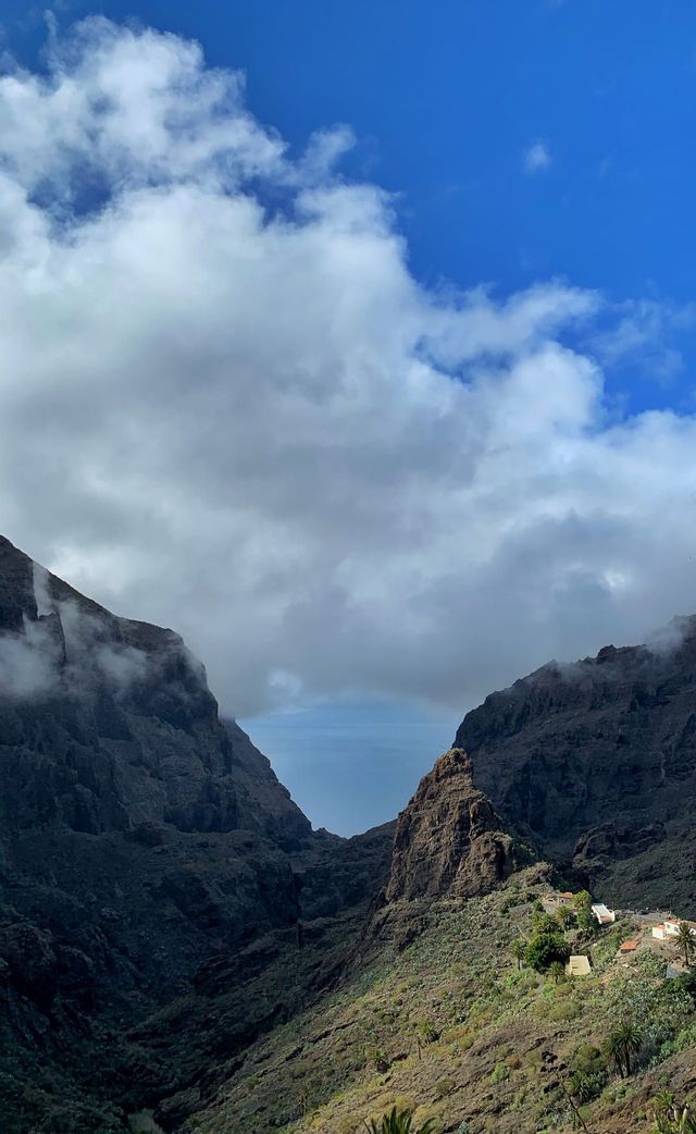 Un valle profundo entre dos escarpadas montañas rocosas se abre hacia el mar, con un pequeño pueblo en una ladera verde bajo un cielo nublado.