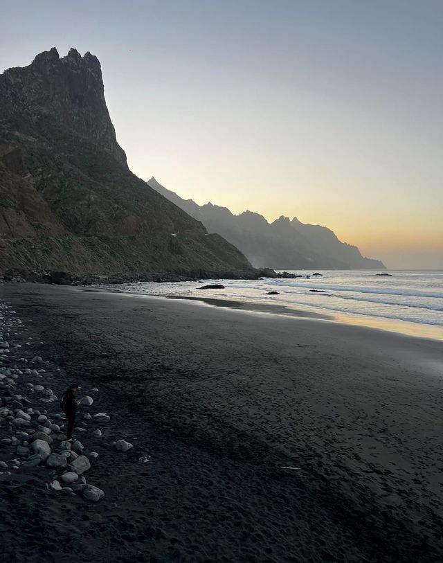 Una persona solitaria se encuentra en una playa de arena negra con rocas, mirando el océano y una escarpada costa montañosa al atardecer.