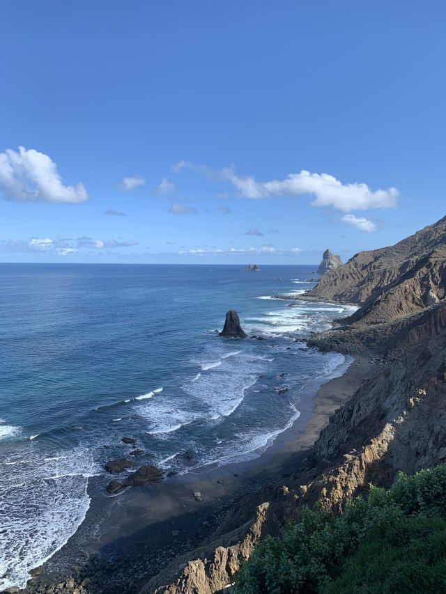 Vista aérea de una costa escarpada con una playa de arena negra, farallones y olas azules del océano bajo un cielo parcialmente nublado.