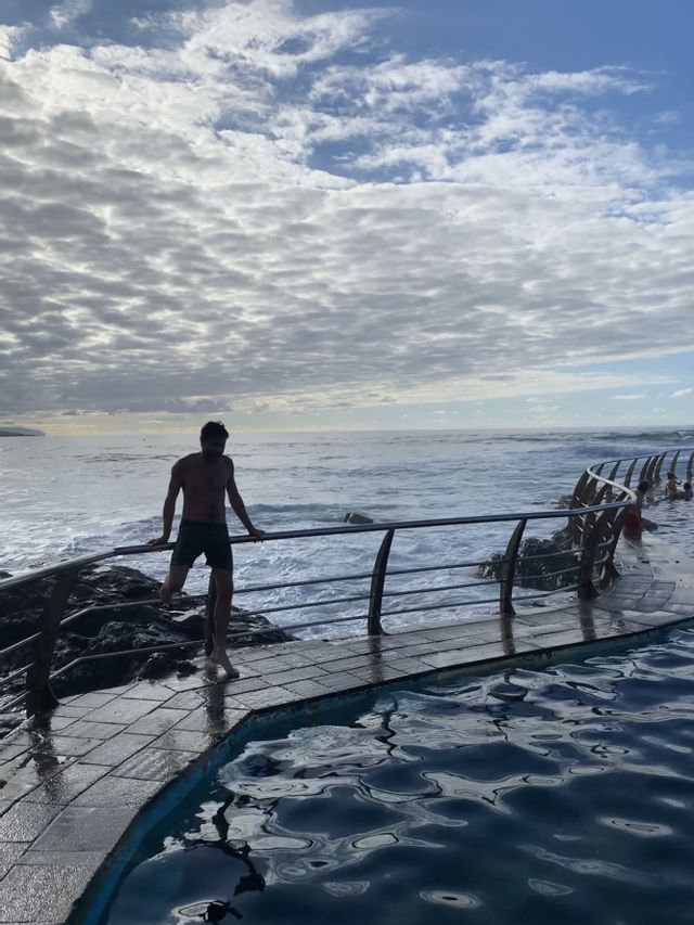Un hombre en pantalones cortos se apoya en la barandilla de una piscina, con el océano y un cielo nublado de fondo.