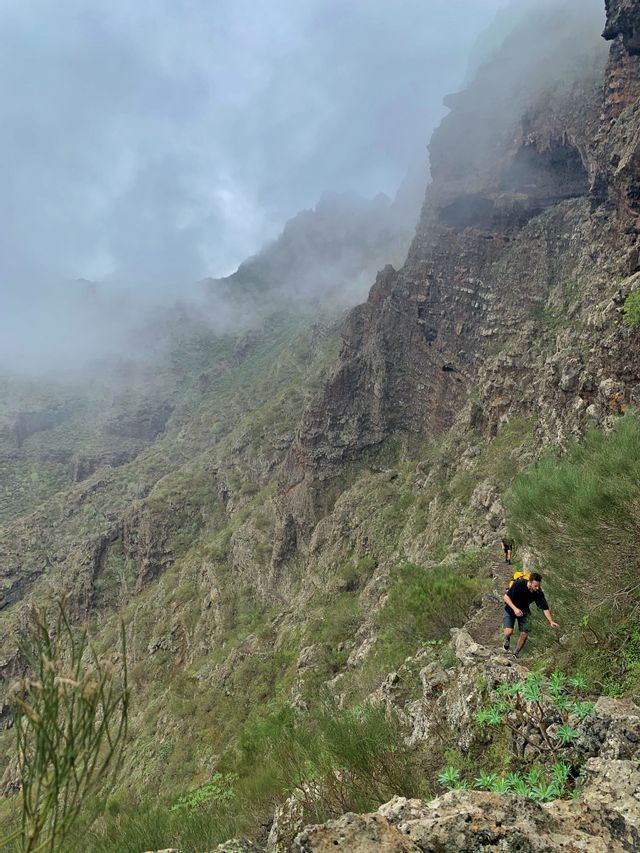 Un viaje en grupo de WeRoad caminando por un sendero angosto en una ladera de montaña verde y empinada, con niebla cubriendo los picos.