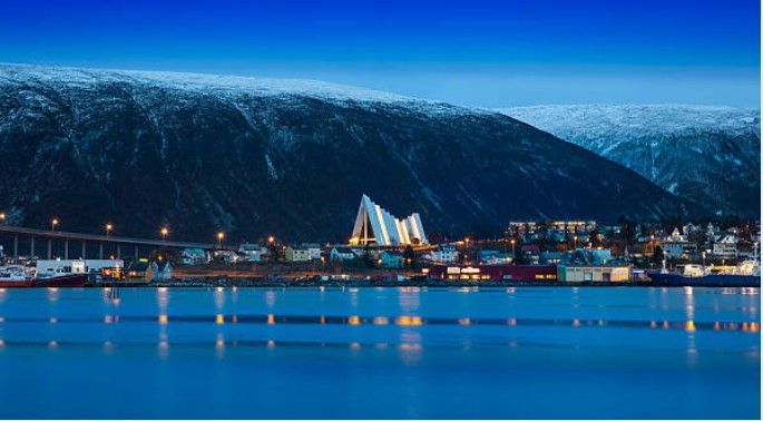 Una città costiera è illuminata al crepuscolo, con luci che si riflettono sull'acqua di fronte a una grande montagna innevata.