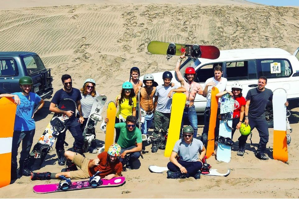 Un groupe WeRoad en voyage, portant des casques, pose avec ses sandboards pour une photo dans un vaste désert de dunes de sable.