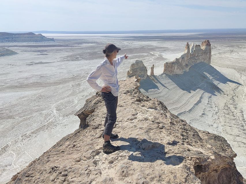 Una persona con gorra se encuentra en una estrecha cresta rocosa, señalando un vasto valle desértico soleado con formaciones rocosas blancas.