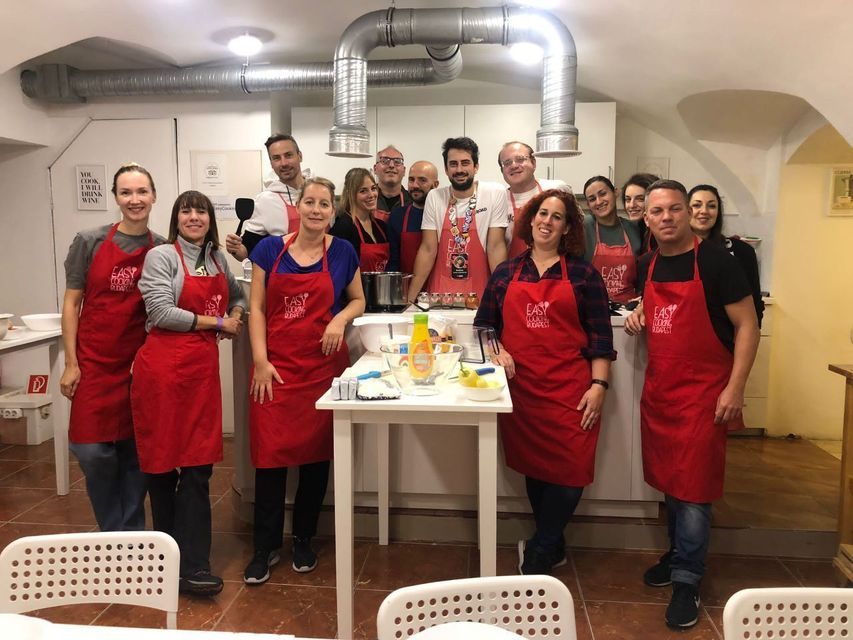 Eine reisende WeRoad-Gruppe in roten Schürzen lächelt für ein Gruppenfoto während eines Kochkurses in einer Küche.