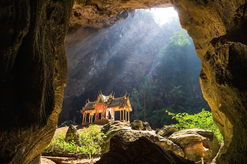 Un padiglione dorato in stile thailandese si trova all'interno di un'ampia grotta, illuminato da un fascio di luce solare proveniente da un'apertura in alto.