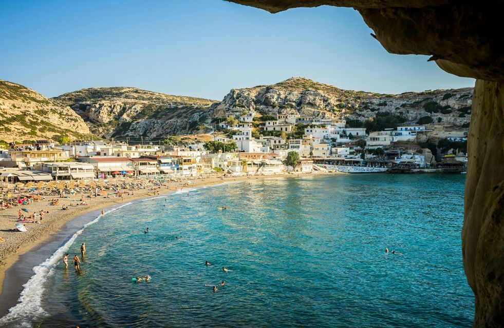 Vista da una grotta su una spiaggia affollata con persone che nuotano in acqua turchese, accanto a un villaggio in collina.