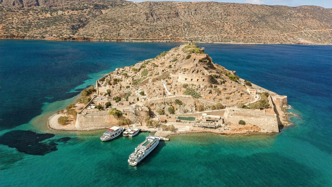 Vista aerea di un'isola storica fortificata con rovine in pietra, con diverse barche ormeggiate nell'acqua turchese circostante.