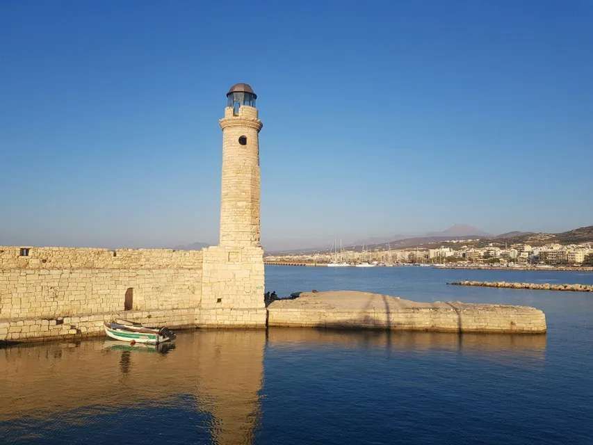 Un faro de piedra se alza al final de un muelle junto a un mar azul en calma, con una ciudad portuaria visible al fondo.