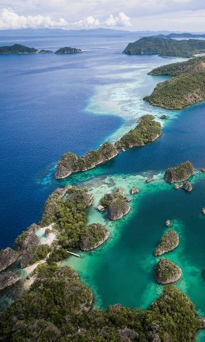 Una vista aérea de un archipiélago tropical con islas verdes exuberantes, agua turquesa cristalina y arrecifes de coral visibles bajo un cielo parcialmente nublado.
