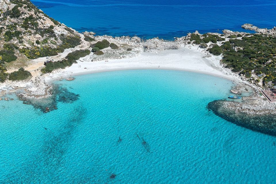 Veduta aerea di una spiaggia di sabbia bianca appartata in una cala, con acqua turchese cristallina che incontra una costa rocciosa.