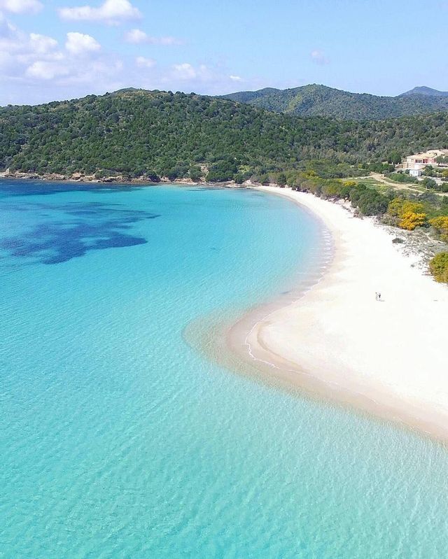 Una vista aerea di una spiaggia di sabbia bianca e curva che incontra acque limpide color turchese, con due persone che camminano vicino a colline verdi.