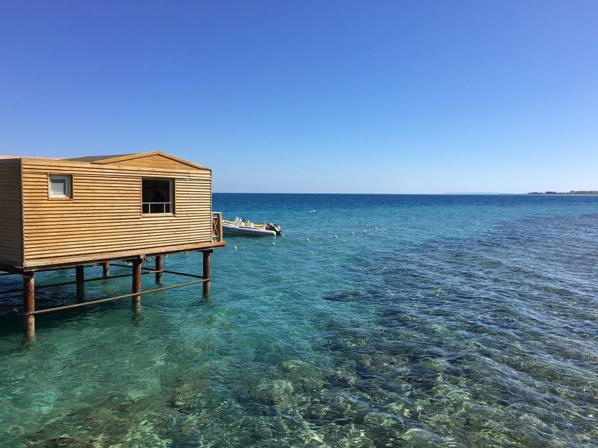 Una capanna di legno su palafitte si erge su un mare cristallino color turchese accanto a una piccola barca ormeggiata sotto un cielo sereno.