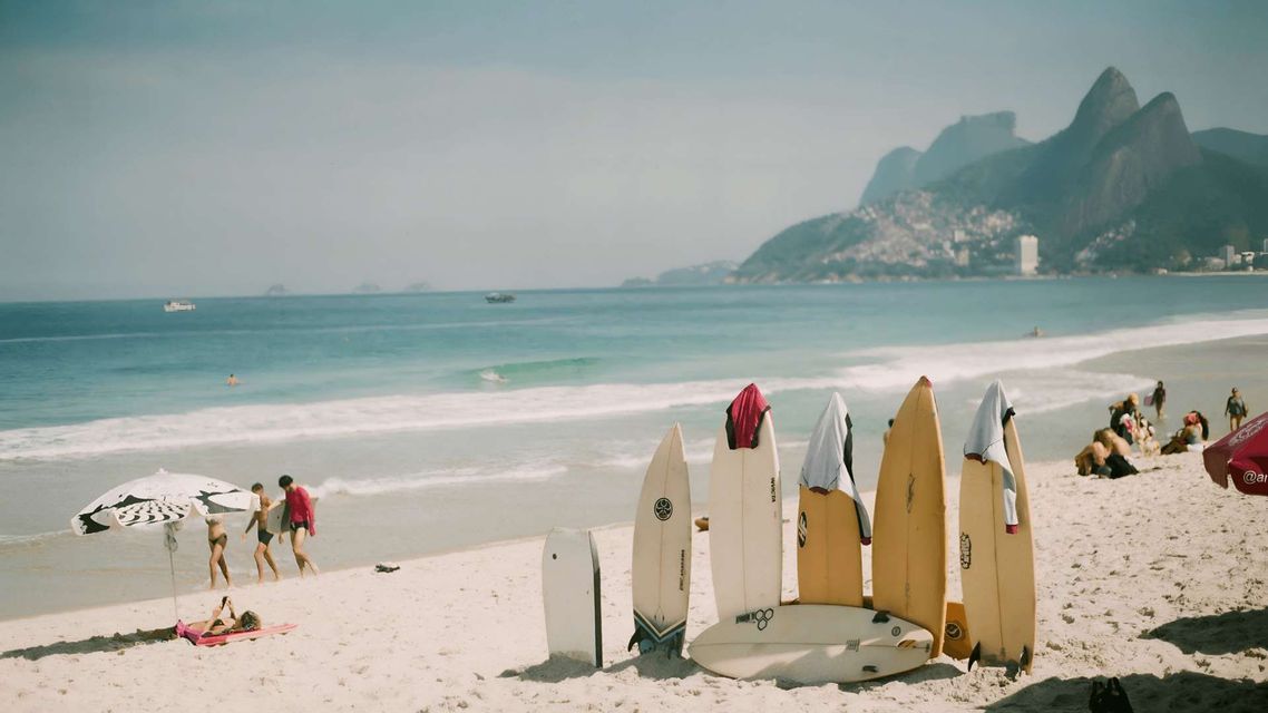 Auf einem sonnigen Strand stehen mehrere Surfbretter aufrecht im Sand. Im Hintergrund entspannen Menschen am Meer vor einer Bergkulisse.