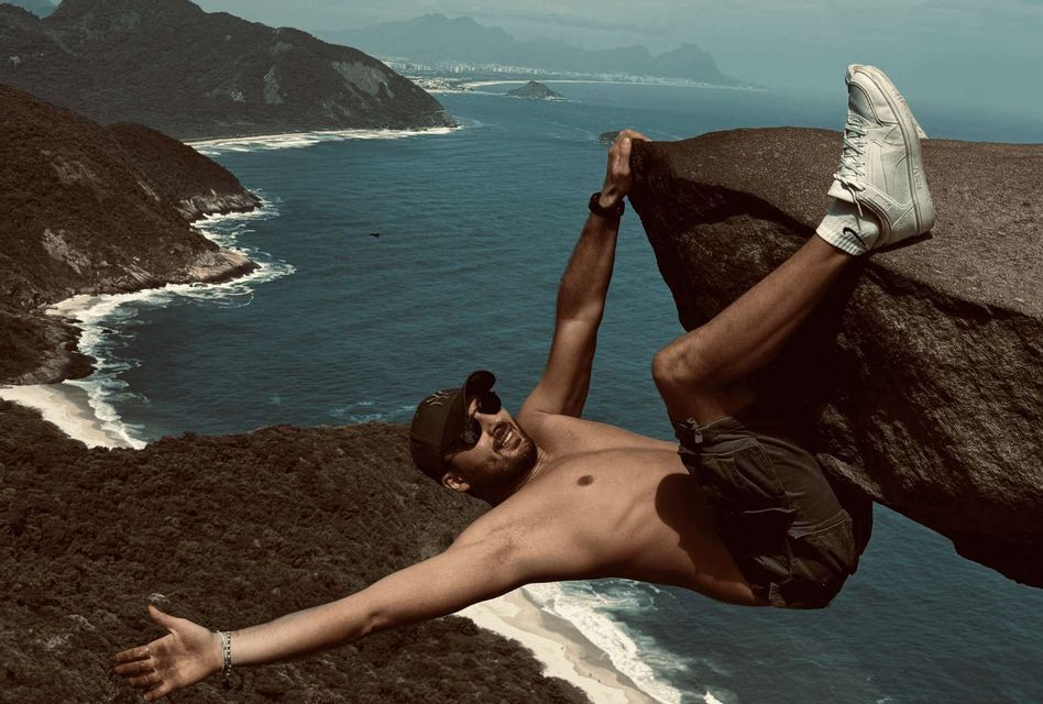 Un uomo a torso nudo con un cappello si appende a una roccia, in posa per una foto su una vista costiera con montagne e una spiaggia sabbiosa.