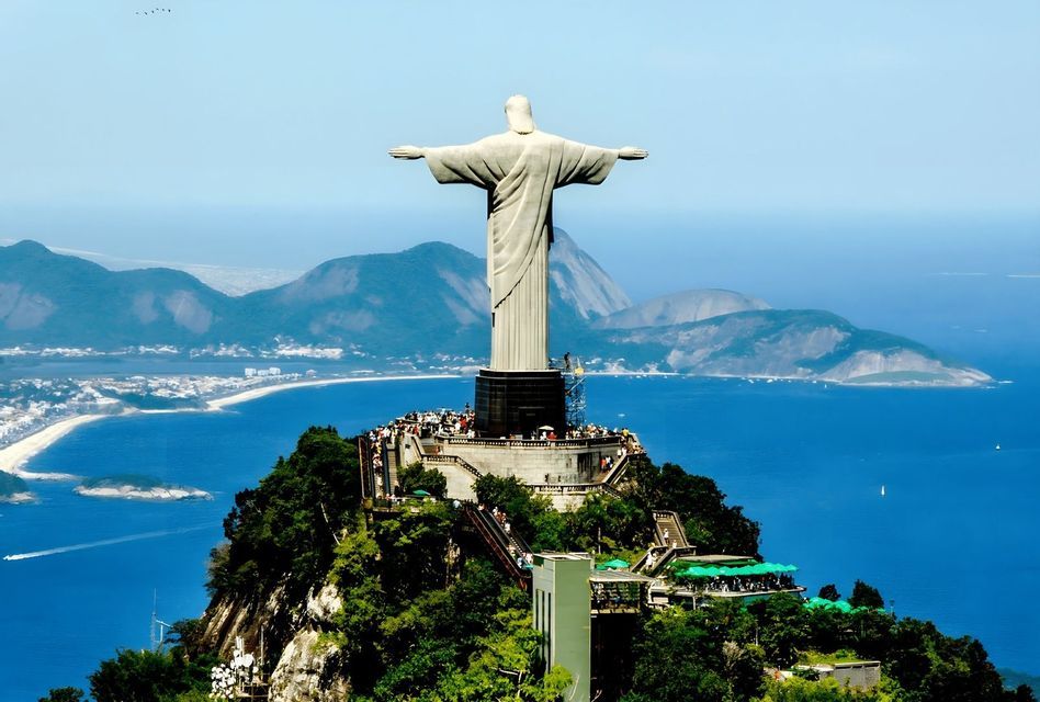 Una grande statua di pietra con le braccia aperte si erge sulla cima di una montagna, affacciata su una piattaforma panoramica affollata, una città costiera e un vasto oceano.