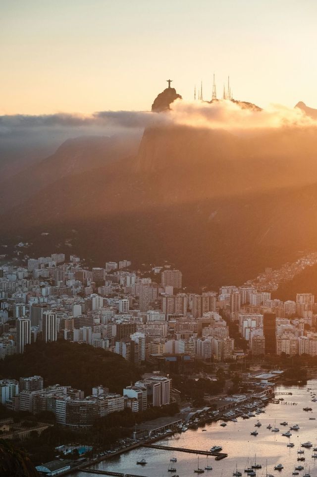 Una vasta città costiera e una baia con barche all'alba, con la sagoma di una grande statua su una cima montuosa distante.