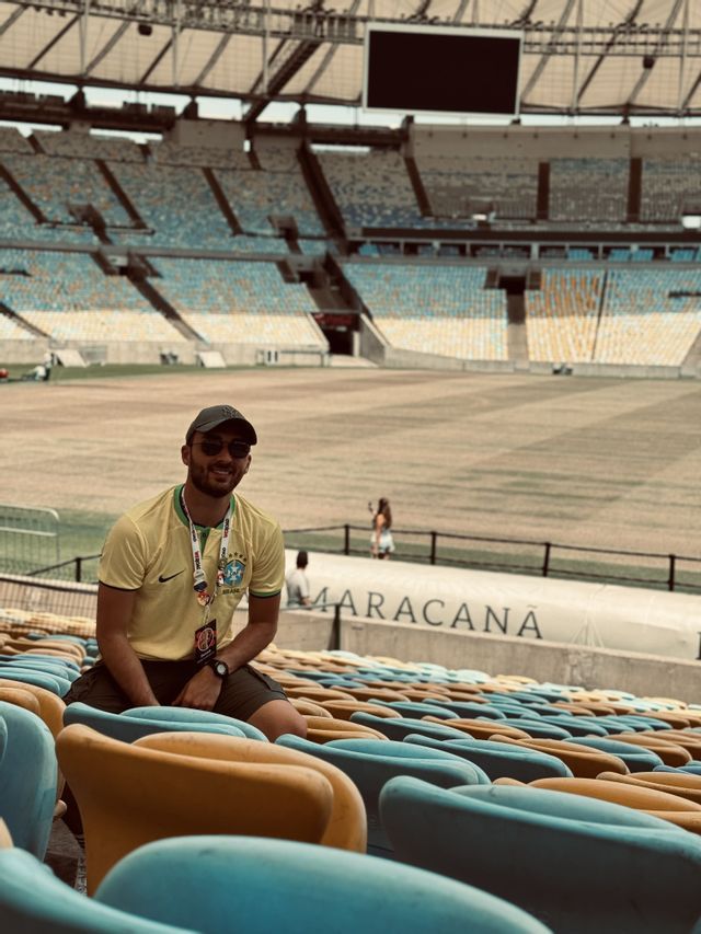 Un uomo sorridente che indossa una maglia gialla e un cappellino siede sulle tribune vuote di un grande stadio.