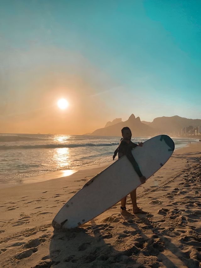 Un surfista in muta tiene una tavola da surf su una spiaggia sabbiosa mentre il sole tramonta sull'oceano e sulle montagne lontane.