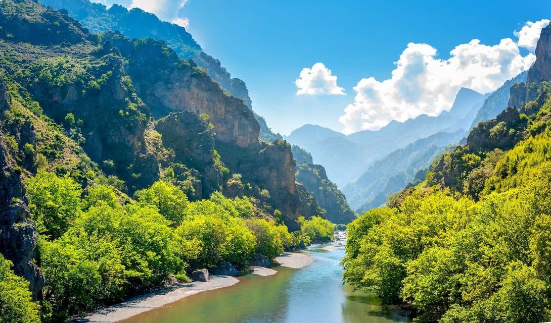 A river winds through a lush green mountain valley with steep cliffs under a blue sky with white clouds.