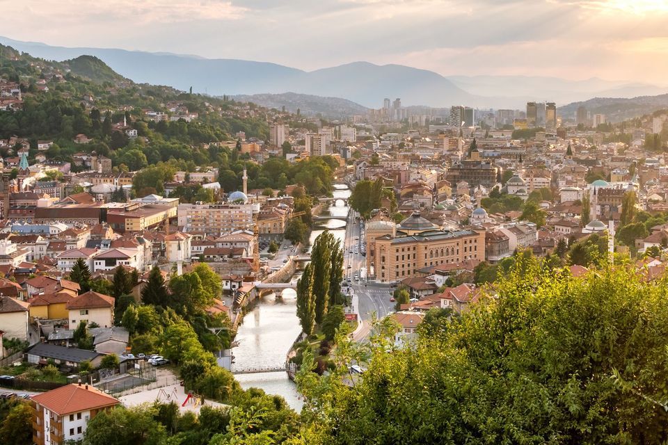 Una vista de la ciudad desde un ángulo alto al atardecer, con un río y varios puentes cruzando el centro y montañas al fondo.
